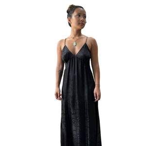 Könnte beinhalten: Ein schwarzes Maxikleid mit Spaghettiträgern und V-Ausschnitt. Das Kleid hat ein dezentes, geprägtes Blumenmuster. Das Model trägt schwarze Mary Jane-Schuhe und eine goldene Halskette mit Anhänger.