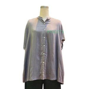 May include: A gray short-sleeved button-down shirt with a mandarin collar. The shirt has white buttons and the word "indochi" in green. The shirt is paired with black pants. The shirt is displayed on a mannequin.