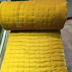 以下が含まれることがあります： 正方形のパターンが施された、黄色のベルベットのキルトの毛布。毛布は上部が丸められており、キルトの質感が見えます。