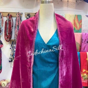 May include: A fuchsia velvet shawl with white stitching around the edges. The shawl is draped over a mannequin wearing a teal blue dress. The text "IndochinaSilk" is visible on the shawl.