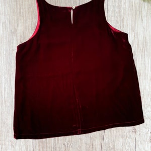 May include: A sleeveless, burgundy velvet top with a keyhole neckline and a small button closure at the back. The top has a soft, luxurious appearance and a slightly flared shape. The interior lining is a contrasting red color.