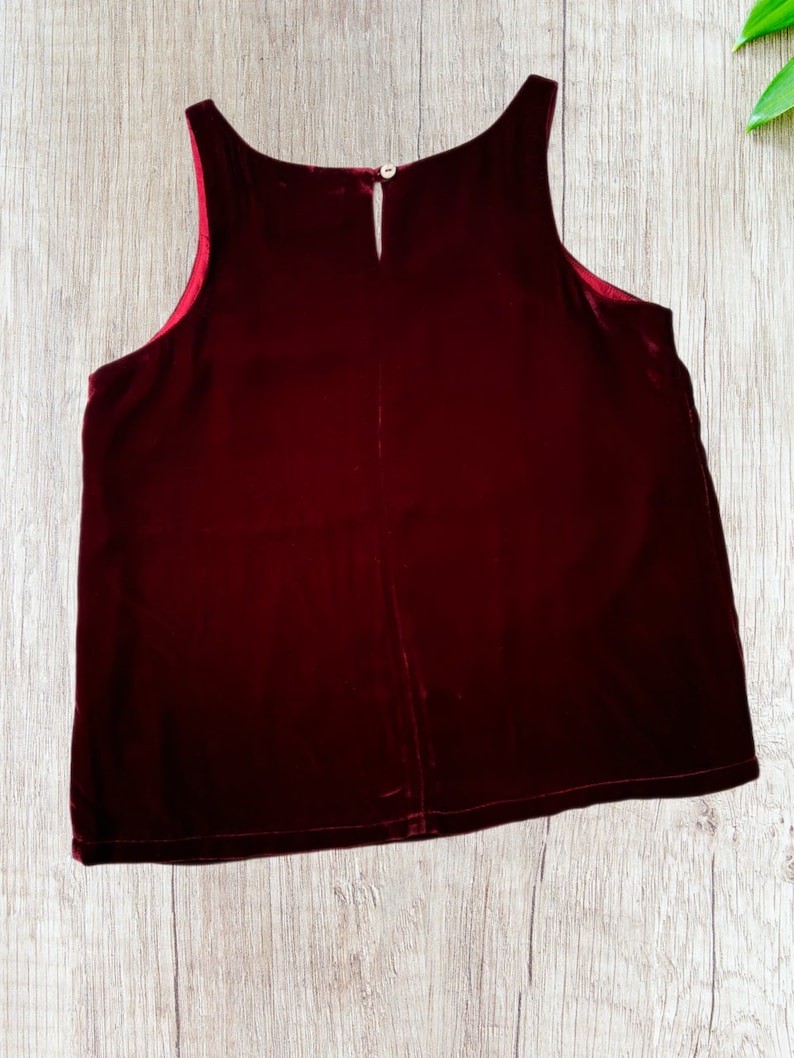 May include: A sleeveless, burgundy velvet top with a keyhole neckline and a small button closure at the back. The top has a soft, luxurious appearance and a slightly flared shape. The interior lining is a contrasting red color.