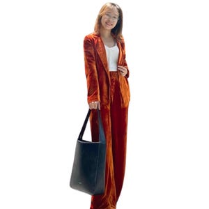 May include: A burnt orange velvet pantsuit with a matching blazer and wide-leg trousers. The outfit is paired with a white top and a black tote bag. The ensemble is a stylish, monochromatic look.