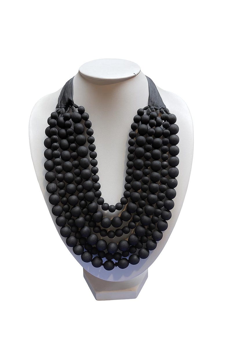 May include: A black beaded necklace displayed on a white mannequin bust. The necklace features multiple strands of matte black beads of varying sizes, creating a layered effect. The necklace is secured with a dark gray ribbon.