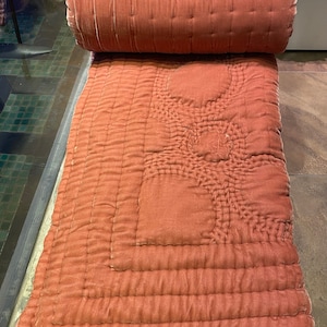 Puede incluir: Una colcha de terciopelo rosa polvoriento enrollada con un patr&oacute;n acolchado. La colcha est&aacute; doblada por la mitad y los bordes son visibles.