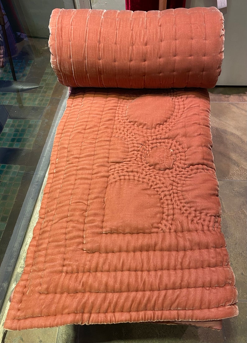 Puede incluir: Una colcha de terciopelo rosa polvoriento enrollada con un patr&oacute;n acolchado. La colcha est&aacute; doblada por la mitad y los bordes son visibles.