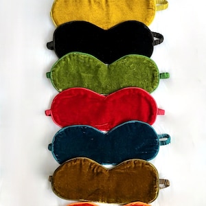 Può includere: Un set di otto maschere per dormire in velluto di vari colori, tra cui blu, giallo, nero, verde, rosso, verde acqua, marrone e arancione. Le maschere sono disposte in una pila, con la maschera blu in cima e la maschera arancione in fondo.