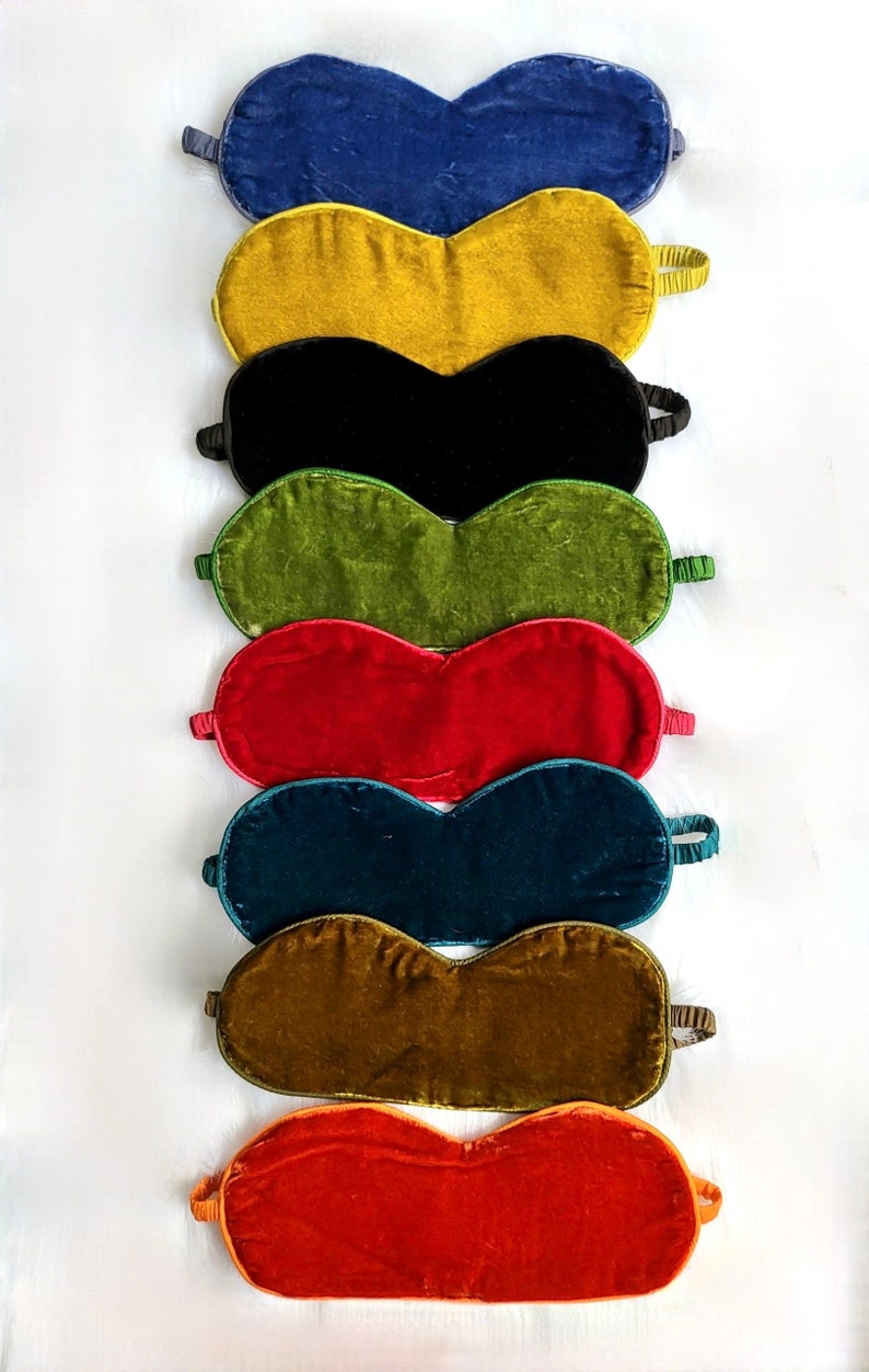 May include: A set of eight velvet sleep masks in various colors, including blue, yellow, black, green, red, teal, brown, and orange. The masks are arranged in a stack, with the blue mask on top and the orange mask on the bottom.