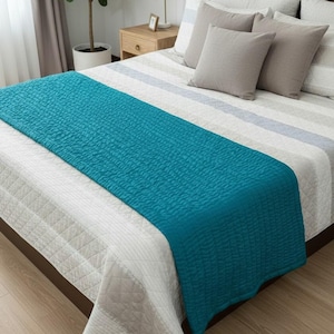 May include: A bed with a white and gray striped comforter and gray pillows. A teal textured bed runner is draped across the bed. A small wooden nightstand with a lamp and a potted plant are visible.