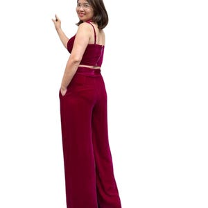 May include: A two-piece burgundy outfit featuring a crop top with spaghetti straps and high-waisted, wide-leg pants. The outfit is made of a soft, velvet-like material. The model is wearing glasses and a necklace.