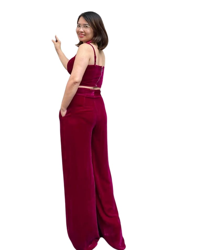 May include: A two-piece burgundy outfit featuring a crop top with spaghetti straps and high-waisted, wide-leg pants. The outfit is made of a soft, velvet-like material. The model is wearing glasses and a necklace.