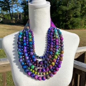 May include: A layered necklace featuring fabric-covered beads in a spectrum of colours, including blue, purple, green, and yellow. The necklace is displayed on a white mannequin. The beads are of varying sizes. The background shows a natural outdoor setting.
