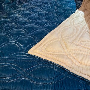 May include: A teal-coloured, quilted blanket with a repeating, scalloped pattern. The blanket is made of a soft, velvety material. A section of a cream-coloured blanket with a stitched, curved design is also visible.