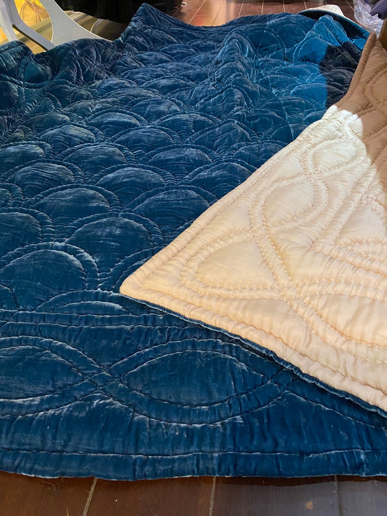 May include: A teal-coloured, quilted blanket with a repeating, scalloped pattern. The blanket is made of a soft, velvety material. A section of a cream-coloured blanket with a stitched, curved design is also visible.