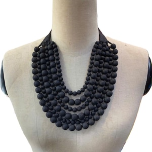 May include: A black beaded necklace displayed on a mannequin. The necklace features multiple strands of matte black beads of varying sizes, creating a layered effect. The beads are strung together, forming a statement piece.