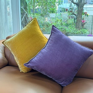 Op de afbeelding: Twee decoratieve kussens, een in mosterdgele fluweel en de andere in paarse fluweel, liggen op een bruine leren bank.