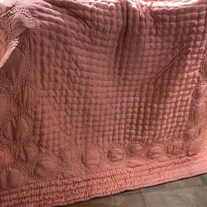 Puede incluir: Una manta acolchada rosa con un patr&oacute;n decorativo de c&iacute;rculos y cuadrados. La manta est&aacute; doblada y puesta sobre un suelo de baldosas.