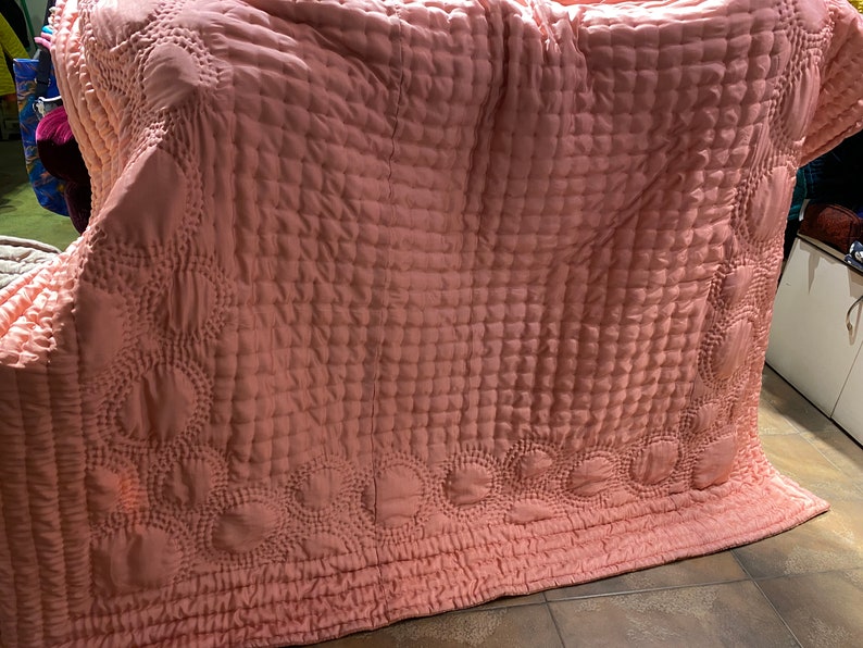 Puede incluir: Una manta acolchada rosa con un patr&oacute;n decorativo de c&iacute;rculos y cuadrados. La manta est&aacute; doblada y puesta sobre un suelo de baldosas.