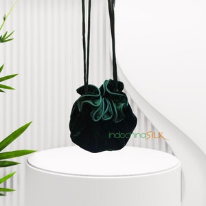 May include: A dark green velvet drawstring bag with a ruffled top. The bag is hanging from two cords and is displayed on a white pedestal.