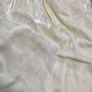 以下が含まれることがあります： 柔らかくビロードのような質感のオフホワイトの生地。生地はドレープされており、その光沢を際立たせる折り目と影を作り出しています。この素材は、ベルベットまたは同様の豪華なテキスタイルのように見えます。
