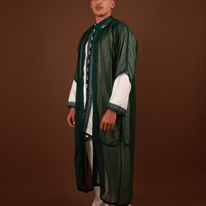 May include: A man wearing a long, dark green, sheer robe with white trim and white pants. He is wearing white leather slippers.