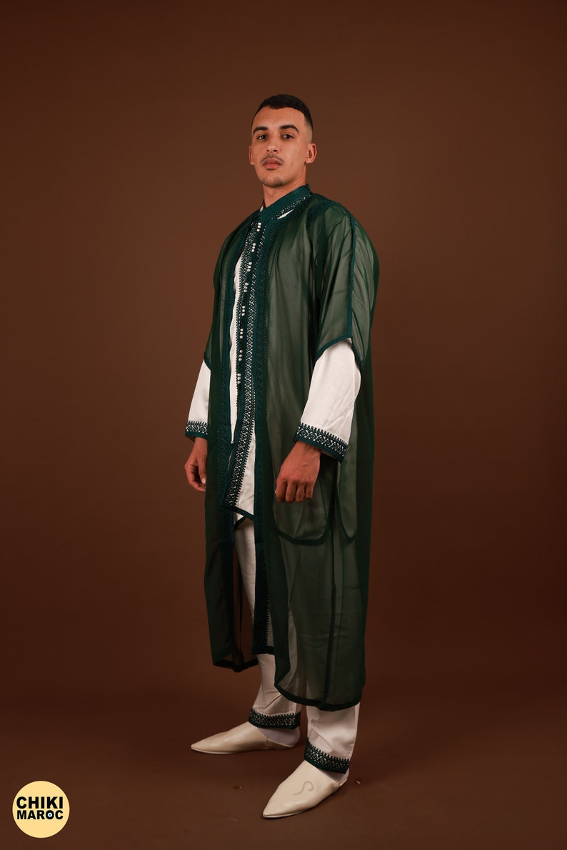 May include: A man wearing a long, dark green, sheer robe with white trim and white pants. He is wearing white leather slippers.