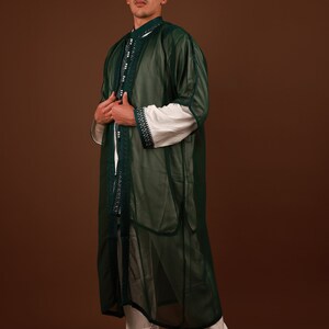May include: A man wearing a long, green, sheer robe with white trim and intricate embroidery. He is also wearing white pants and white leather shoes.
