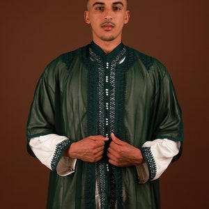May include: A man wearing a long, dark green robe with intricate white embroidery and pearl-like embellishments. The robe has a high collar and is open in the front, revealing a white undershirt. The man is standing against a brown background.