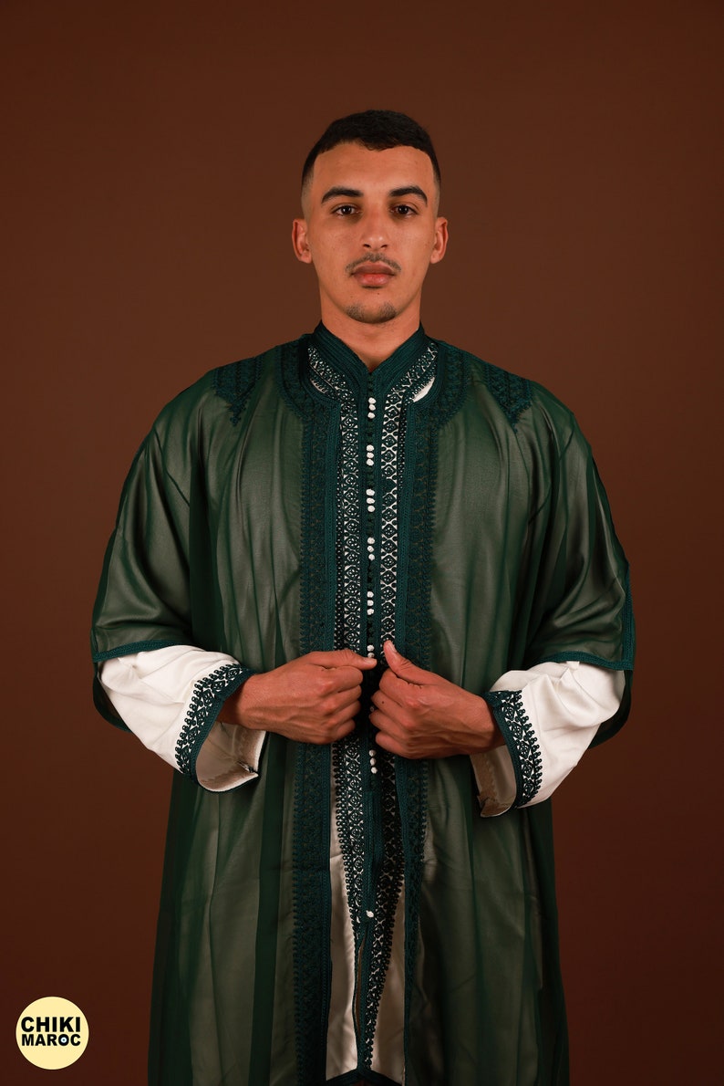 May include: A man wearing a long, dark green robe with intricate white embroidery and pearl-like embellishments. The robe has a high collar and is open in the front, revealing a white undershirt. The man is standing against a brown background.