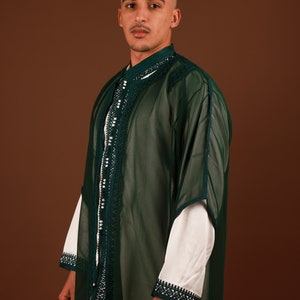 May include: A man wearing a long, green, sheer robe with white trim and pearl-like embellishments. The robe has a high collar and long sleeves. The man is standing against a brown background.