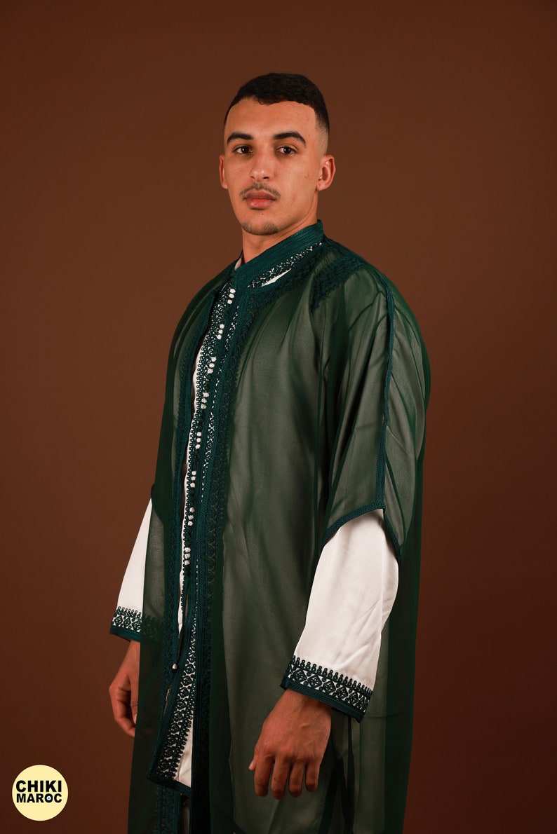 May include: A man wearing a long, green, sheer robe with white trim and pearl-like embellishments. The robe has a high collar and long sleeves. The man is standing against a brown background.