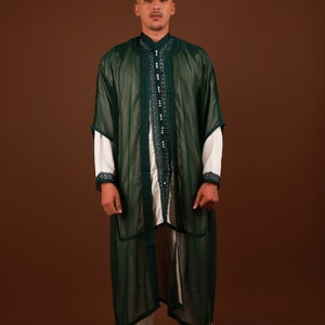 May include: A man wearing a long, green, sheer robe with white trim and intricate embroidery. The robe is open in the front, revealing a white undershirt and white pants with matching trim. The man is standing on a brown background.
