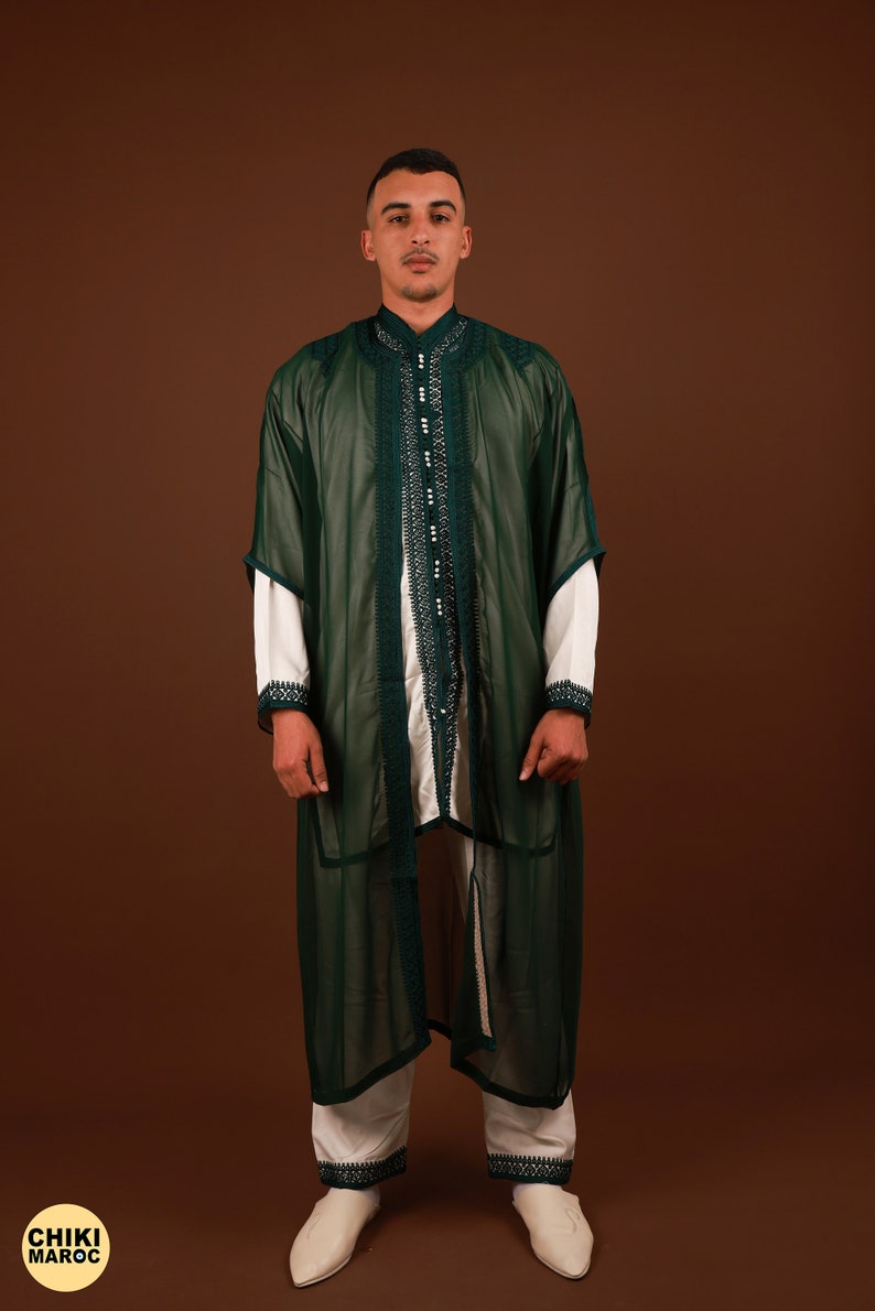 May include: A man wearing a long, green, sheer robe with white trim and intricate embroidery. The robe is open in the front, revealing a white undershirt and white pants with matching trim. The man is standing on a brown background.