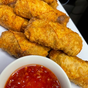 Può includere: Un piatto bianco con una varietà di involtini primavera croccanti dorati. Un piccolo piatto bianco pieno di una salsa rossa per intingere si trova in primo piano.