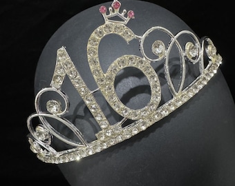 Könnte beinhalten: Silberfarbene Tiara mit der Zahl "16", besetzt mit klaren Strasssteinen. Eine kleine Krone mit rosa Akzenten sitzt auf der "16". Die Tiara wird auf einem schwarzen Hintergrund präsentiert, was einen Kontrast erzeugt. Ideal für eine sechzehnte Geburtstagsfeier.