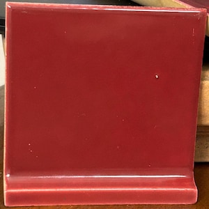 May include: A square, glossy, red ceramic tile with a rounded edge. The tile has a smooth, reflective surface and a deep, rich color. The tile is likely for use in a kitchen or bathroom.