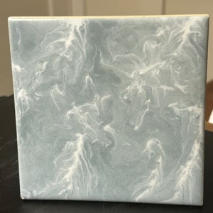 May include: Square ceramic tile with a marbled design. The tile features a light gray background with swirling white patterns, creating a flowing, abstract effect. The edges are smooth and the overall aesthetic is modern and decorative.