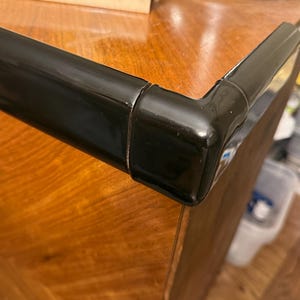 May include: Close-up of a black, glossy corner trim piece. The trim is attached to a wooden surface with a warm, reddish-brown grain. A light-colored wooden piece is visible in the background, along with a portion of a record player.