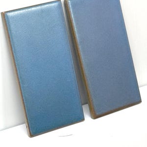 May include: Two rectangular, blue ceramic tiles with a glossy finish and brown edges. The tiles are leaning against each other, showcasing their color and texture. The tiles are likely for home decor or construction.