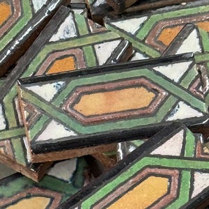 May include: A collection of rectangular ceramic tiles featuring a geometric pattern with a central ochre hexagon surrounded by green, white, and brown lines. The tiles have a glossy finish and dark edges, arranged in a stacked, overlapping pile.