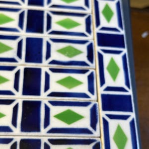 May include: A collection of rectangular ceramic tiles arranged in a grid pattern. Each tile features a white background with a dark blue border and a centered green diamond shape. The tiles are laid out in rows and columns with visible grout lines.