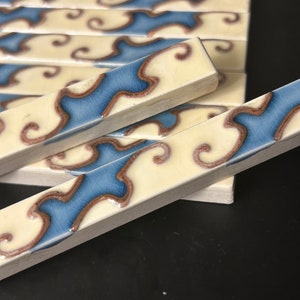 May include: Rectangular ceramic tiles with a cream-colored background and a blue and brown decorative pattern. The tiles are arranged in a stack, with some tiles in the foreground. The design features a wavy blue stripe with brown swirls.