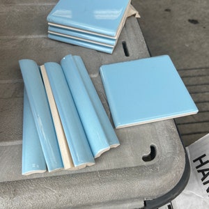 May include: A collection of light blue ceramic tiles, including a stack of square tiles and several elongated, curved edge trim pieces. Each piece features a smooth, glossy finish with a consistent pale blue color and a light-colored base.