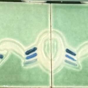 May include: A series of four square ceramic tiles arranged horizontally, featuring a glossy light green glaze. Each tile displays a continuous embossed wave pattern in cream, accented with three parallel dark blue and light blue stripes.