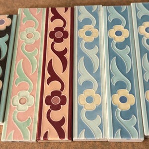 May include: Six rectangular ceramic border tiles arranged side by side. Each tile features a raised relief pattern of repeating four-petaled flowers connected by scrolling vines. The tiles display various color combinations including black, pink, maroon, and blue.