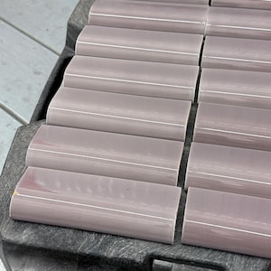 May include: A collection of mauve-colored, rectangular tiles with a curved top edge and a white accent line. The tiles are arranged in rows on a dark gray surface, likely for a construction or design project.