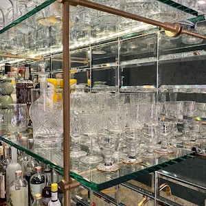 Może przedstawiać: Szafka barowa z lustrem, z szklanymi p&oacute;łkami i elementami mosiężnymi. P&oacute;łki są wypełnione szkłem, w tym kieliszkami do wina, kieliszkami do koktajli i kieliszkami do shot&oacute;w.