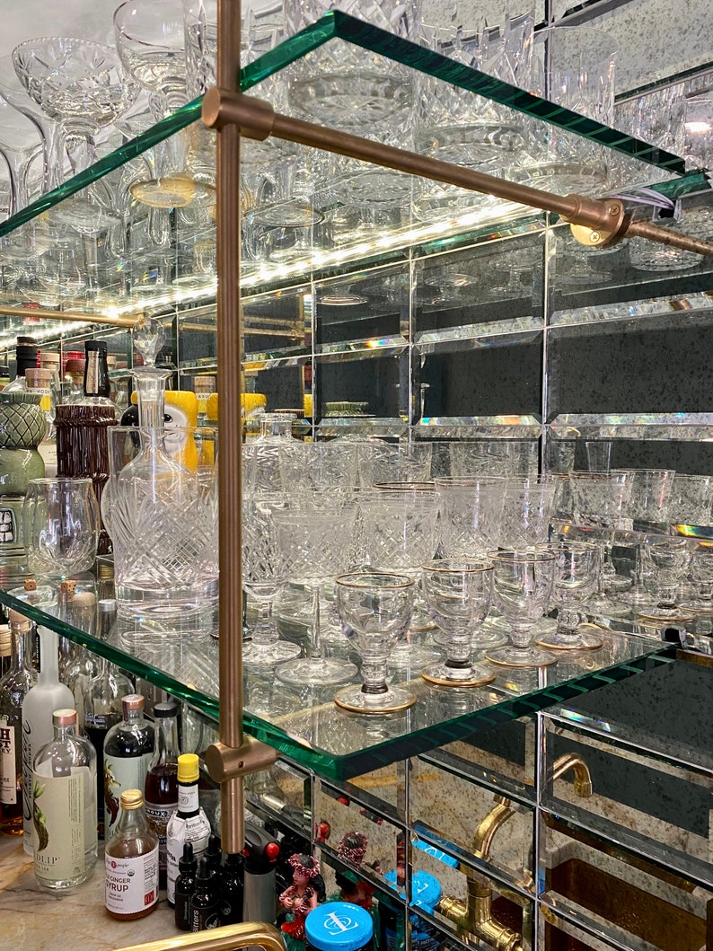 Może przedstawiać: Szafka barowa z lustrem, z szklanymi p&oacute;łkami i elementami mosiężnymi. P&oacute;łki są wypełnione szkłem, w tym kieliszkami do wina, kieliszkami do koktajli i kieliszkami do shot&oacute;w.