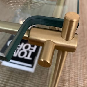 Może przedstawiać: Zbliżenie na złoty metalowy łącznik łączący dwa kawałki szkła. Łącznik ma kształt litery T i jest wypolerowany do błyszczącego wykończenia.