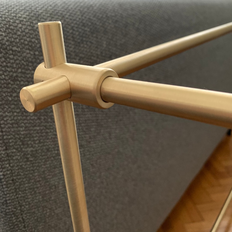 Może przedstawiać: Zbliżenie na złoty metalowy wspornik p&oacute;łki z okrągłym złączem. Wspornik wykonany jest z gładkiego, polerowanego metalu i ma prosty, nowoczesny design.